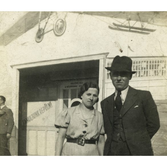Elegáns pár egy épület előtt, „Tanuljunk repülni” felirat repülőgép rajzával, jármű, közlekedés, Horthy-korszak, 1936. 1930-as évek, Eredeti fotó, papírkép. 